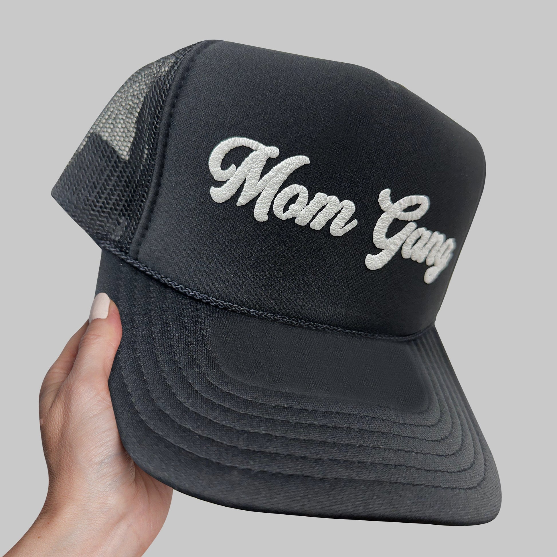 a hand holding a black trucker hat with the word mom gay on it