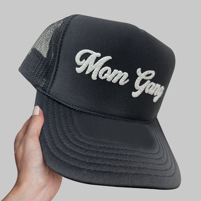 a hand holding a black trucker hat with the word mom gay on it