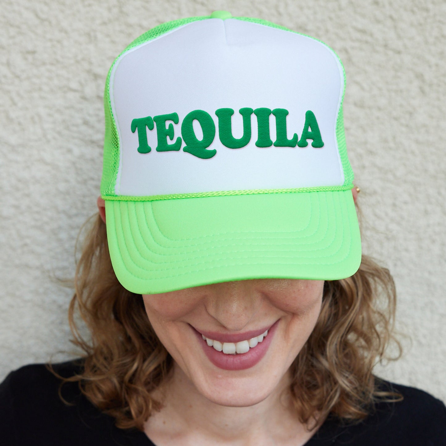 a smiling woman wearing a green and white tequila trucker hat